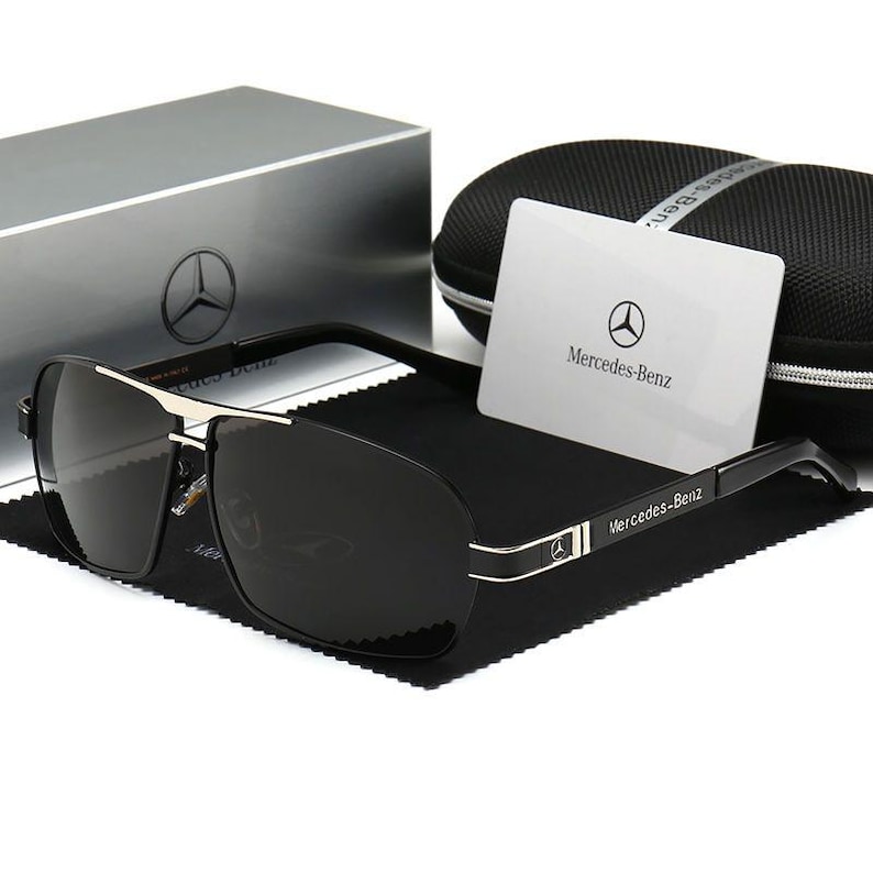 May include: Black sunglasses with a silver frame and dark lenses. The sunglasses are displayed with a case, cleaning cloth, and a card that says "Mercedes-Benz". The sunglasses have the Mercedes-Benz logo on the frame.