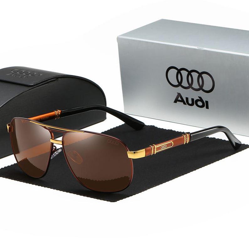 Op de afbeelding: Aviator zonnebril met bruine glazen en een goudkleurig montuur. De pootjes zijn zwart met bruine en gouden accenten. Het Audi-logo is zichtbaar op de etui en de doos op de achtergrond.