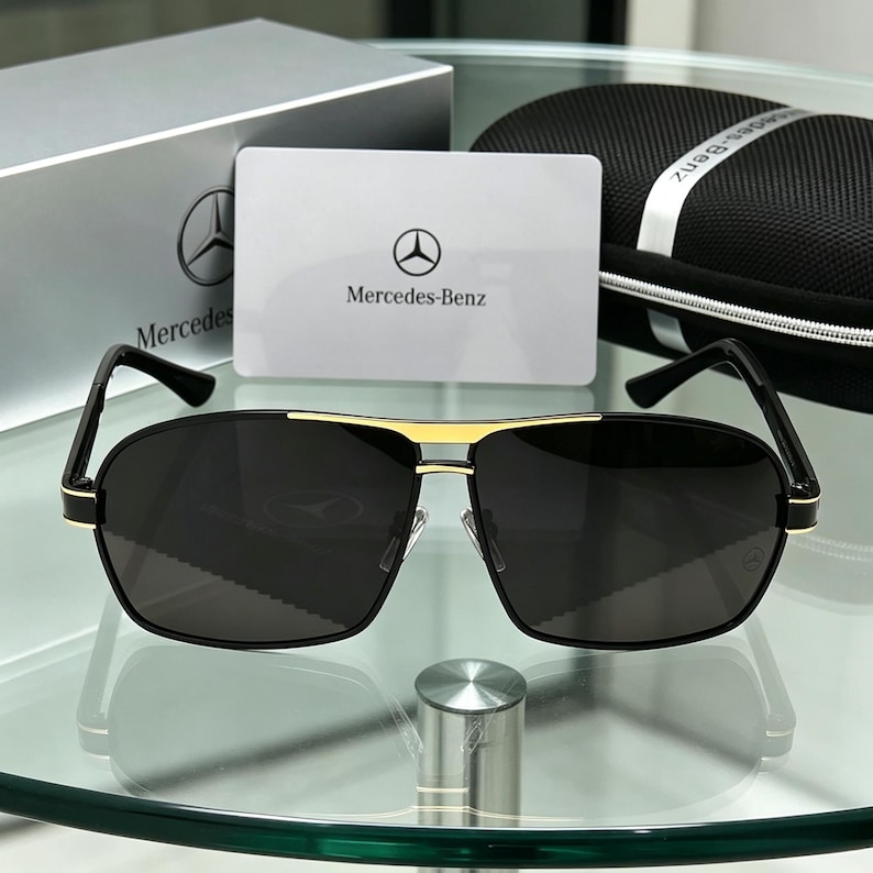May include: Black sunglasses with a gold bridge and accents, featuring the Mercedes-Benz logo on the lens and a matching case. The sunglasses are displayed with a white card that has the Mercedes-Benz logo and text.