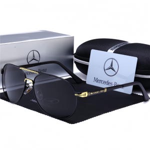 Op de afbeelding: Zwarte piloten zonnebril met gouden details en het Mercedes-Benz logo op de pootjes. De afbeelding toont een etui, een schoonmaakdoekje en een kaart met het Mercedes-Benz embleem.