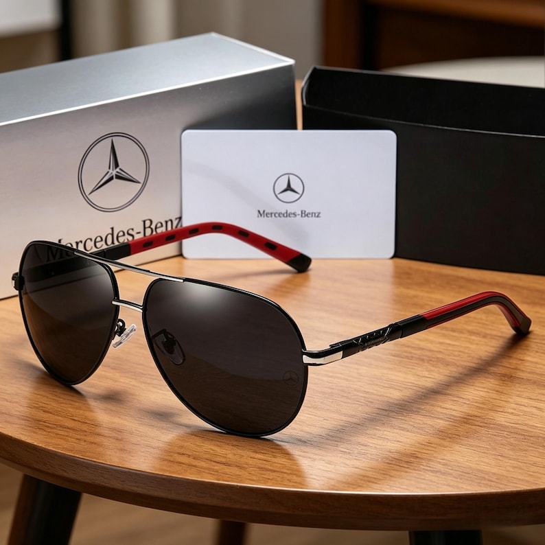 May include: Black aviator sunglasses with a silver frame and red and black temples. The sunglasses are displayed with a box and card, both featuring the Mercedes-Benz logo.