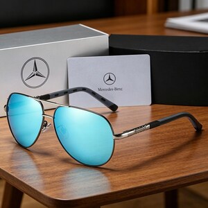 May include: Aviator-style sunglasses with blue mirrored lenses and silver frames. The temples are black and gray. The sunglasses are displayed on a wooden surface, with a Mercedes-Benz logo on a card and box in the background.