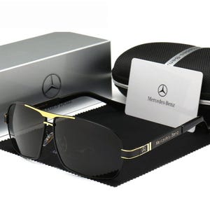 May include: Black sunglasses with gold detailing and the Mercedes-Benz logo on the frame and card. The sunglasses are presented with a case and box, both featuring the Mercedes-Benz emblem.