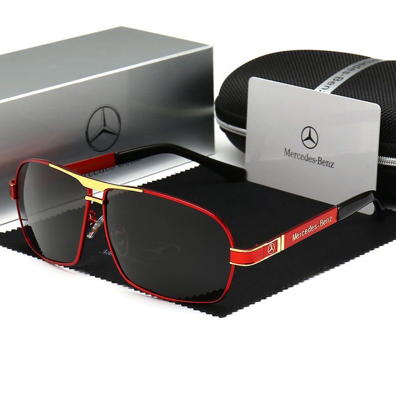May include: Red and gold sunglasses with dark lenses. The sunglasses have a rectangular shape and a gold bridge. The temples are black with red accents and the Mercedes-Benz logo. A silver box and a black case are in the background.