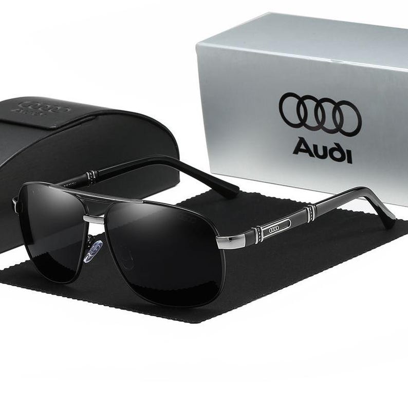 Op de afbeelding: Zwarte piloten zonnebril met een zilverkleurig montuur en zwarte pootjes. De zonnebril wordt gepresenteerd met een zwart etui en een zilveren doos met het Audi-logo.