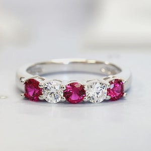 Op de afbeelding: Een zilverkleurige ring met vijf rond geslepen stenen op een rij. De stenen wisselen af tussen levendig roze en helder wit, vastgehouden door metalen pootjes op een gladde, gepolijste band die om de vinger sluit.