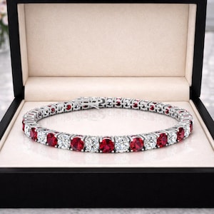 May include: A tennis bracelet featuring alternating round-cut red rubies and clear white diamonds set in a polished silver-toned metal band. The bracelet is displayed in an open black jewelry box with a cream-colored interior lining.