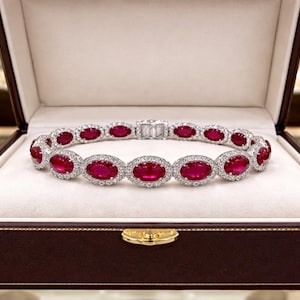 May include: A bracelet featuring a series of oval-cut vibrant red gemstones, each surrounded by a halo of small white stones. The piece is set in a metallic frame and is displayed inside a brown rectangular jewelry box with a cream-colored interior.