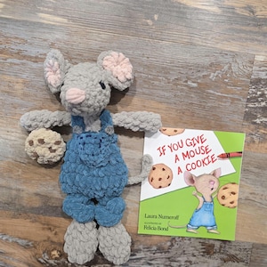 Puede incluir: Un ratón de ganchillo gris hecho a mano con un peto azul, sosteniendo una galleta, junto a una copia del libro infantil "Si le das una galleta a un ratón". El ratón tiene detalles rosas en las orejas y la nariz.