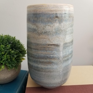 May include: A cylindrical ceramic vase featuring a reactive glaze with horizontal bands of muted blue, soft gray, and earthy brown tones. The surface has a subtle, textured appearance with blended color transitions and a smooth, rounded rim.