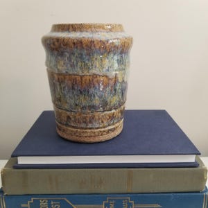 May include: A cylindrical ceramic vase featuring a reactive glaze with blended tones of brown, tan, blue, and grey. The vessel has a slightly flared rim, a smooth upper body, and a textured, unglazed base that shows the natural clay color.