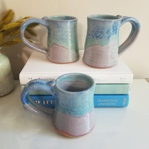 May include: Three ceramic mugs with a glossy glaze featuring a color gradient of light blue, turquoise, and soft lavender. Each mug has a sturdy handle and a smooth, rounded rim. The mugs are arranged with two sitting atop a stack of books.