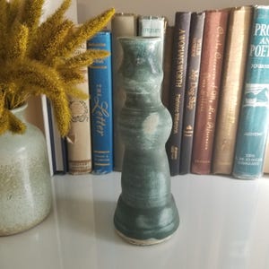 May include: A tall, slender ceramic vase with a matte, mottled forest green glaze. The vase features a flared base, a rounded midsection, and a narrow neck with a slightly wider rim. The surface shows subtle variations in color and texture.