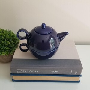 May include: A dark blue ceramic tea for one set consisting of a rounded teapot with a matching lid and a cup that nests directly underneath the teapot. The teapot features a curved handle and a short spout, while the cup has a circular handle.