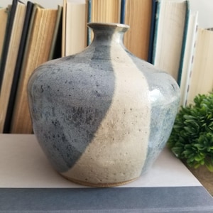 May include: A ceramic vase with a rounded body and a narrow neck, featuring a two-tone glaze pattern of mottled slate gray and textured off-white. The surface has a visible speckled finish and a smooth, glossy appearance across the entire exterior.
