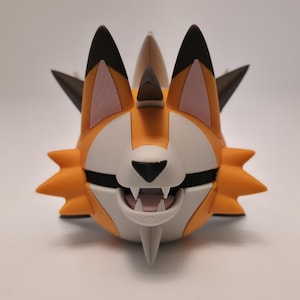 May include: A stylized orange and white creature figurine with pointed ears, dark brown tips, and a central horn. The character features a wide, open mouth revealing small white teeth, a black nose, and several sharp, angular protrusions extending from the sides.