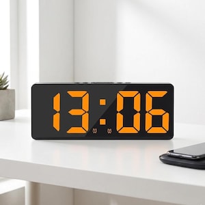 May include: A black digital alarm clock showing the time 13:06 in orange numerals. The rectangular clock is on a white surface. A smartphone is placed on a wireless charging pad next to the clock.
