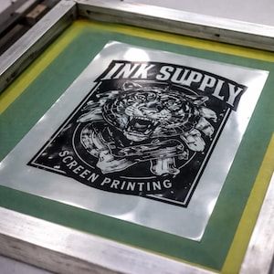 Könnte beinhalten: Ein Siebdruckrahmen aus Metall mit gelbem Siebgewebe und einer transparenten Filmvorlage, die eine schwarz-weiße Grafik eines brüllenden Tigers zeigt. Der Text INK SUPPLY steht über dem Tiger und SCREEN PRINTING steht darunter.