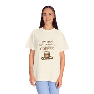 May include: Cream-colored t-shirt with the text "MY DOG NEEDS SPACE AND I NEED COFFEE" in brown. A graphic of a coffee cup with a paw print and a leash is below the text. The shirt is made of a soft, comfortable material.