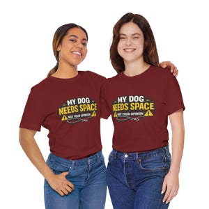 May include: Two people wearing matching maroon t-shirts with the text "MY DOG NEEDS SPACE, NOT YOUR OPINION" in yellow and white. The shirts have a graphic of a leash and a caution sign. They are wearing blue jeans.