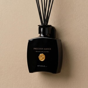 Op de afbeelding: Een zwarte diffuserfles met de tekst "Precious Amber" en "Private Collection" in het wit. De fles heeft een gouden zegel en het woord "Rituals..." in het wit. Zwarte rietjes zijn in de bovenkant van de fles gestoken.