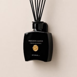 Op de afbeelding: Een zwarte glazen fles voor een geurstokjesverspreider met een gouden rond embleem op de voorzijde. De tekst luidt Precious Amber, Private Collection en Rituals. Meerdere dunne zwarte stokjes steken uit de glanzende fles naar boven.