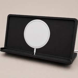 Può includere: Un supporto di ricarica rettangolare nero con un caricabatterie wireless magnetico bianco circolare centrato. Il supporto presenta un bordo orizzontale alla base per sostenere un dispositivo e un cavo di alimentazione bianco.