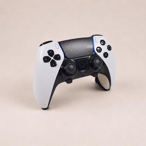 Peut inclure: Une manette de jeu vidéo sans fil blanche et noire dotée d'un pavé tactile central texturé, de deux joysticks analogiques noirs, d'une croix directionnelle et de quatre boutons d'action sur la droite. L'appareil inclut une icône de micro.