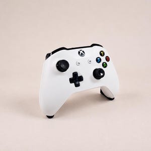 Op de afbeelding: Een witte draadloze videogamecontroller met een zwarte richtingspad, twee zwarte analoge sticks en vier actieknoppen in de kleuren blauw, geel, rood en groen. Het midden van de controller toont het zwart-witte Xbox-logo.