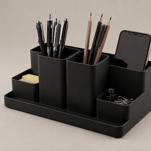 Op de afbeelding: Een matzwarte bureau-organizer met meerdere vakken van verschillende hoogtes. Het bevat diverse pennen, potloden, een smartphone, een stapel gele memoblaadjes en zilverkleurige metalen klemmen. De structuur is rechthoekig met getrapte secties.