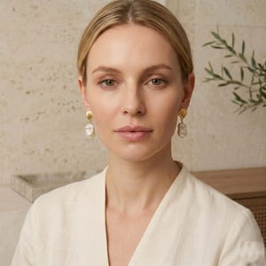 May include: Gold-coloured earrings featuring a seashell design and large, irregular white pearls. The earrings are dangling and worn by a person with blonde hair and a cream-coloured top. The background is a neutral beige.