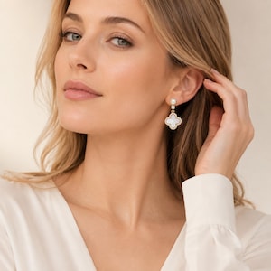 Peut inclure: Boucles d'oreilles dorées avec un pendentif blanc en forme de trèfle et une perle. Les boucles d'oreilles ont un design délicat. Les boucles d'oreilles sont un bijou classique.