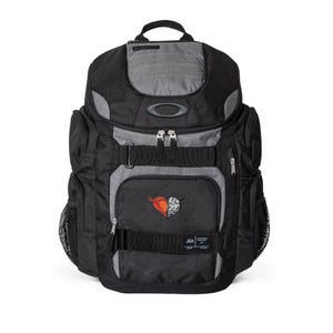 May include: Black and gray backpack with multiple compartments, featuring a top handle and adjustable straps. The front pocket displays a heart-shaped design with orange and white details. The Oakley logo is visible.