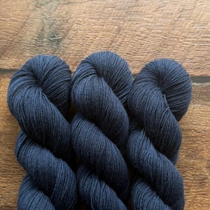 May include: Three skeins of dark navy blue yarn twisted into a tight, uniform spiral. The yarn features a smooth, matte texture with visible individual strands tightly plied together, creating a consistent deep blue color throughout each skein.