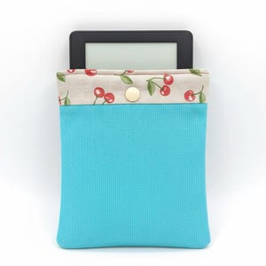 Könnte beinhalten: Eine türkisfarbene E-Reader-Hülle mit einem Kirschdruck-Oberteil. Die Hülle hat einen Knopfverschluss und ist für einen E-Reader konzipiert. Die Oberseite der Hülle hat einen beigen Stoff mit roten Kirschen und grünen Blättern.