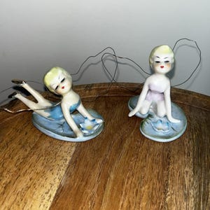 May include: Two porcelain figurines of women in a reclining pose. The figurines have blonde hair, red lips, and are dressed in blue and lavender. They are resting on light blue lily pads with white and yellow flowers. The figurines are on a wooden surface.