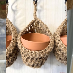 May include: Three hanging woven jute plant holders with terracotta pots. The holders are suspended from black metal hooks. The jute has a natural, light brown color, and the pots are a warm orange-brown.