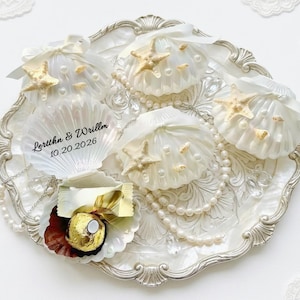 Può includere: Un vassoio decorativo con bordo argentato contiene diversi contenitori bianchi a forma di conchiglia, ciascuno adornato con una stella marina e perle. Il vassoio presenta anche una collana di perle e una conchiglia con il testo "Lertthn & Wrillm 10.20.2026".