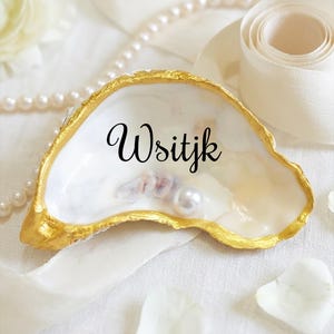 Puede incluir: Un cuenco decorativo de concha de ostra con borde dorado y la palabra "Wsitjk" en escritura negra. La concha contiene una perla y se encuentra sobre tela blanca, con perlas y cinta en el fondo.