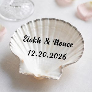 May include: A white scallop shell with a metallic silver rim, featuring the black printed text "Eiokh & Nonce" and the date "12.20.2026" centered on the interior surface of the shell, which has a natural ribbed texture and a glossy finish.