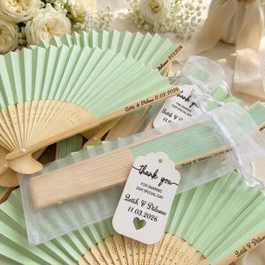 May include: A collection of mint green and natural wood folding fans, some open and some closed. Several fans have text printed on them, and some are packaged with tags that say "thank you" and the date.