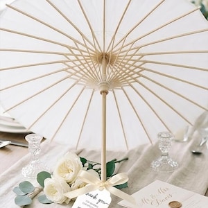 K&ouml;nnte beinhalten: Ein wei&szlig;er Papiersonnenschirm mit einem hellen Holzgestell und Griff. Ein cremefarbenes Band ist um den Griff gebunden und h&auml;lt ein wei&szlig;es Etikett mit der Aufschrift: Thank you for sharing our special day, Mr & Mrs Snmish, 12.16.2026, sowie einem kleinen Herzsymbol.