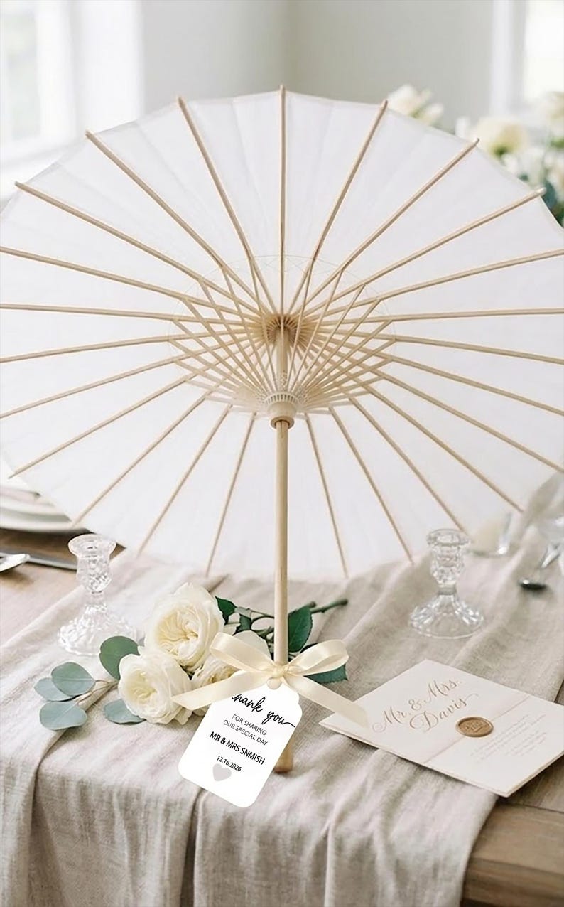 K&ouml;nnte beinhalten: Ein wei&szlig;er Papiersonnenschirm mit einem hellen Holzgestell und Griff. Ein cremefarbenes Band ist um den Griff gebunden und h&auml;lt ein wei&szlig;es Etikett mit der Aufschrift: Thank you for sharing our special day, Mr & Mrs Snmish, 12.16.2026, sowie einem kleinen Herzsymbol.
