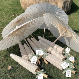 May include: Several off-white paper parasols with wooden handles, some open and some closed, on green grass. Each closed parasol has a tag tied with a green ribbon. A large rock is in the background.