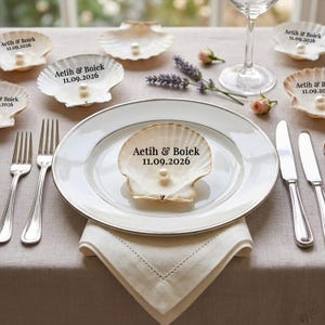 May include: Elegant table setting with white plates, silverware, and seashell place cards. Each seashell holds a pearl and features the text "Aetih & Boiek 11.09.2026". The table is set with a linen napkin and a wine glass.