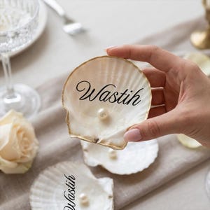 May include: A decorative white seashell with a gold edge, featuring a pearl and the name "Wastih" in black script. Other seashells with pearls are present, indicating a decorative purpose. The setting is on a neutral surface.