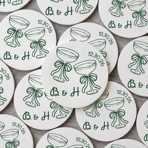 May include: A collection of round white ceramic coasters featuring a dark green line art illustration of two champagne glasses tied with bows. Each coaster is printed with the initials B & H and the date 12.30.26 in a dark green font.