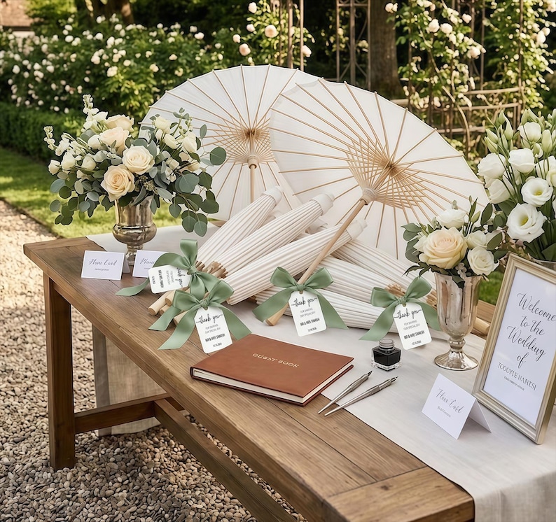 K&ouml;nnte beinhalten: Ein Holztisch zeigt wei&szlig;e Papier-Sonnenschirme mit Bambusrippen, die jeweils mit einem salbeigr&uuml;nen Band und einem Dankesch&ouml;n-Anh&auml;nger versehen sind. Zwei silberne Vasen enthalten cremefarbene Rosen und Eukalyptus sowie ein G&auml;stebuch.