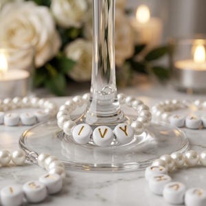 Pode incluir: Encantos de taças de vinho com contas de pérolas brancas que soletram "IVY" com letras douradas. Os encantos estão em uma superfície de mármore, com rosas brancas e velas acesas ao fundo. Outros encantos soletram nomes.