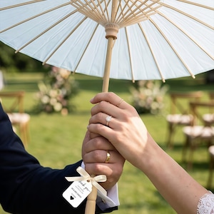 K&ouml;nnte beinhalten: Ein hellblauer Papiersonnenschirm mit Holzgriff und Holzstreben, an dem ein kleines wei&szlig;es Schild mit einem Band befestigt ist. Auf dem Schild steht: Thank you for sharing our special day, Mr & Mrs Samish, 12.16.2026, mit einem schwarzen Herz.